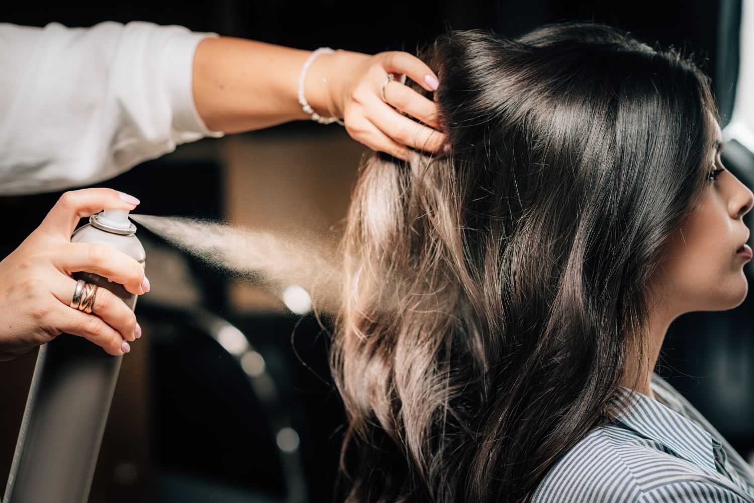 Hair stylist applying spray to woman's long, styled hair in a salon setting.